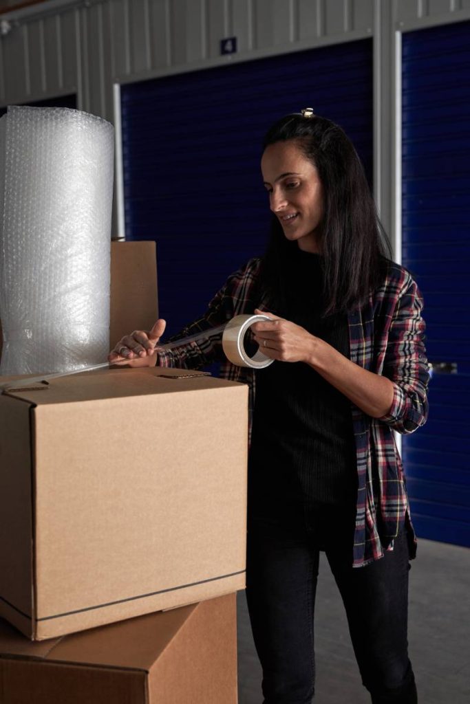 Content female in casual wear packing carton box with belongings using adhesive scotch tape neat self storage during moving day