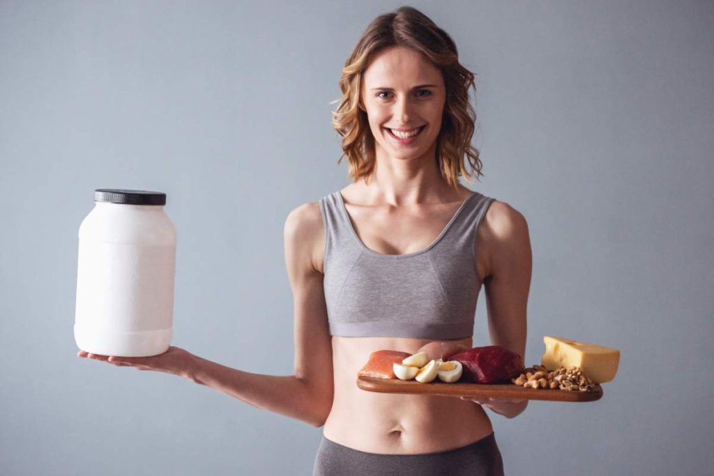 Beautiful young sportswoman is holding a bottle with sport nutrition and a wooden tray with healthy food, on gray background